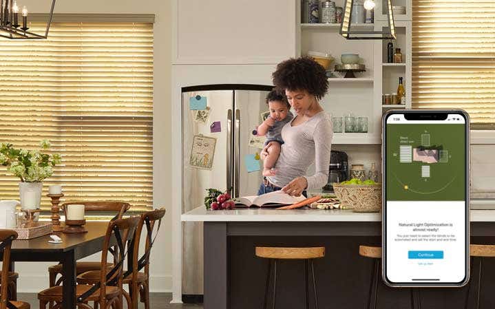 Mom with baby on her hip in the kitchen looking at a book, smart phone on the right of the screen zoomed in showing how smart blinds work