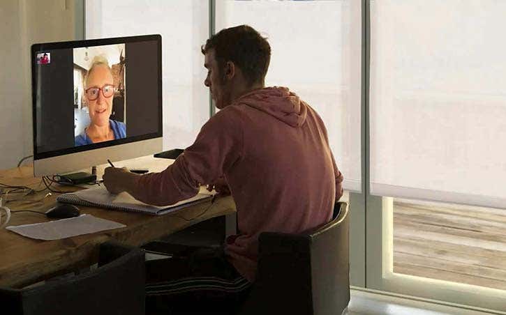 Man on zoom, working with someone and there are white smart roller shades in the windows behind him