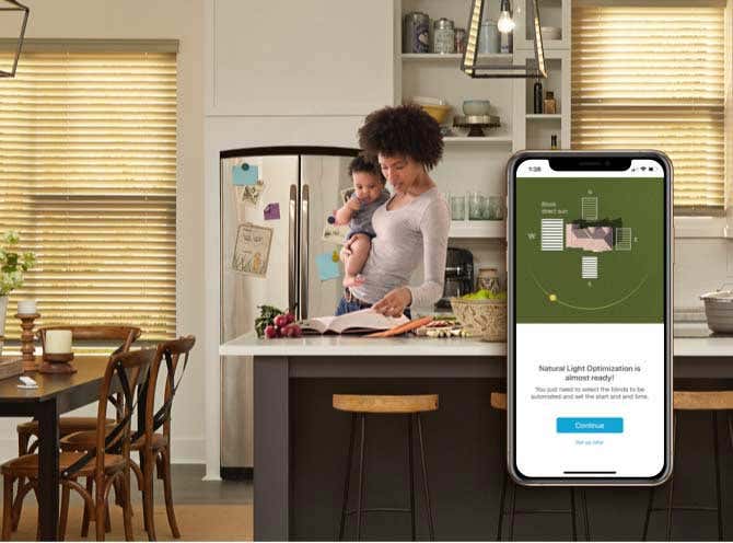 Mom with baby on her hip in the kitchen looking at a book, smart phone on the right of the screen zoomed in showing how smart blinds work